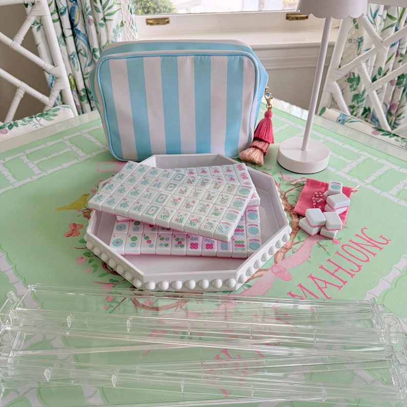 May include: A white and light blue striped cosmetic bag with a pink tassel sits next to a white tray holding a Mahjong set. The tiles are white with floral designs. A small pink bag and dice are also present.