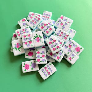 May include: A collection of white Mahjong tiles with pink and purple floral designs. The tiles are rectangular and feature numbers and flower patterns. The background is a solid green color.