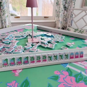 May include: A Mahjong set with white tiles featuring floral and architectural designs, scattered on a green floral patterned surface. The set includes a pink lamp and white trays. The game is set on a table with a green and pink floral design.