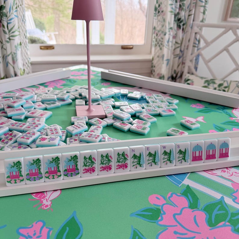 May include: A Mahjong set with white tiles featuring floral and architectural designs, scattered on a green floral patterned surface. The set includes a pink lamp and white trays. The game is set on a table with a green and pink floral design.
