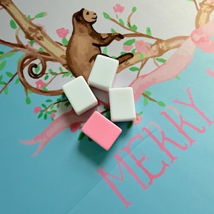 Könnte beinhalten: Vier rechteckige Radiergummis, drei weiße und einer rosa, sind auf einer hellblauen Oberfläche mit einem rosa Blumendesign und dem Wort "MERRY" in Rosa angeordnet. Ein brauner Affe sitzt auf einem Ast.