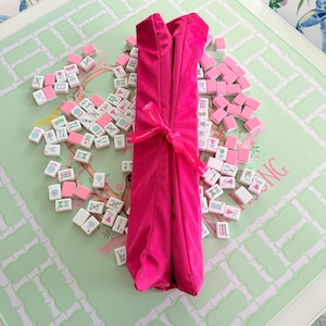 Puede incluir: Un rollo de tela fucsia brillante atado con una cinta a juego, colocado verticalmente sobre una colecci&oacute;n de fichas de mahjong blancas y rosas esparcidas. La cinta presenta un peque&ntilde;o texto impreso y las fichas muestran varios s&iacute;mbolos verdes y rosas.