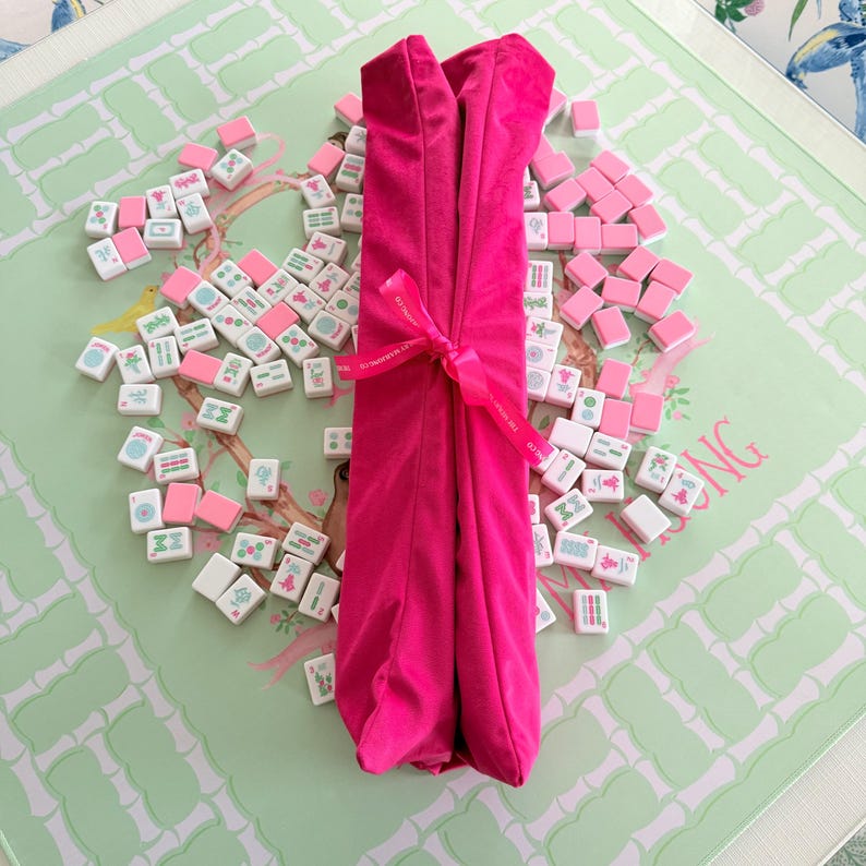 Puede incluir: Un rollo de tela fucsia brillante atado con una cinta a juego, colocado verticalmente sobre una colecci&oacute;n de fichas de mahjong blancas y rosas esparcidas. La cinta presenta un peque&ntilde;o texto impreso y las fichas muestran varios s&iacute;mbolos verdes y rosas.