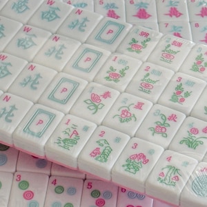 May include: Close-up of a set of white Mahjong tiles with various symbols and characters in pink and teal. Some tiles have floral designs and numbers. The tiles are arranged in a curved pattern.