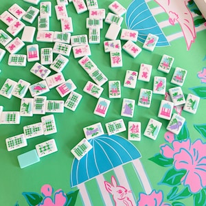 May include: A collection of white and green Mahjong tiles scattered on a green surface with a pink and blue floral pattern. The tiles feature various symbols and characters. A light blue tile is also present. The background includes a pink bunny and a blue umbrella design.