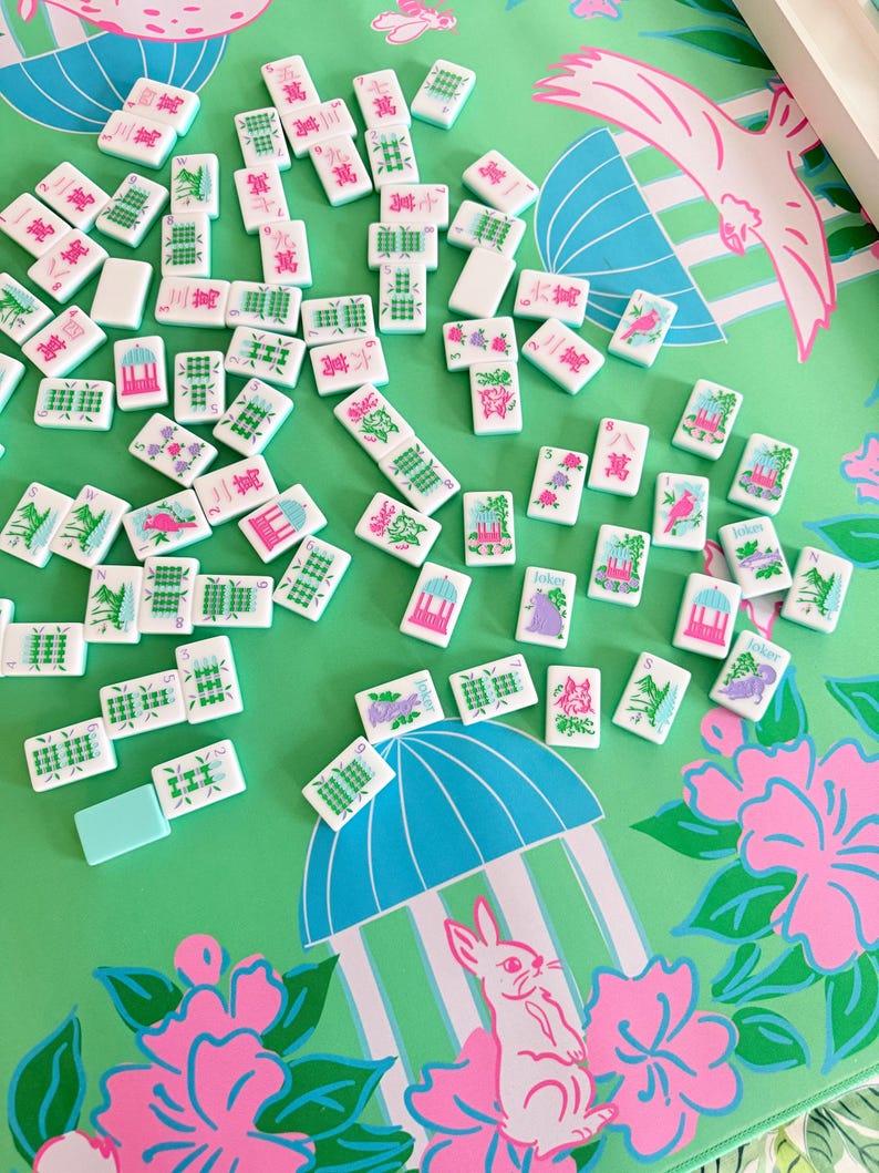 May include: A collection of white and green Mahjong tiles scattered on a green surface with a pink and blue floral pattern. The tiles feature various symbols and characters. A light blue tile is also present. The background includes a pink bunny and a blue umbrella design.