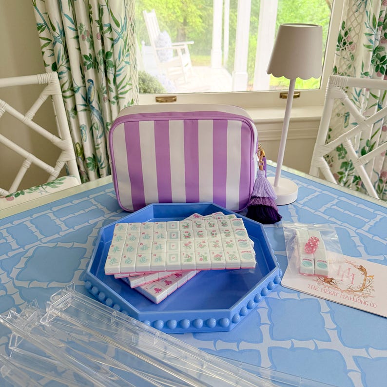 May include: A light blue table displays a Mahjong set with tiles, a striped cosmetic bag, and a blue tray. The Mahjong tiles are white with floral designs. The bag has vertical purple and white stripes. A lamp and accessories are also present.
