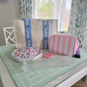 May include: A collection of Mahjong game accessories, including a cream-colored tote bag with blue straps and the word "Mahj" printed on them. Also, a pink and white striped cosmetic bag, a tray of tiles, and a clear acrylic tile rack.