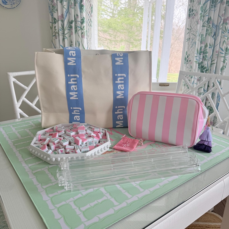 May include: A collection of Mahjong game accessories, including a cream-colored tote bag with blue straps and the word "Mahj" printed on them. Also, a pink and white striped cosmetic bag, a tray of tiles, and a clear acrylic tile rack.