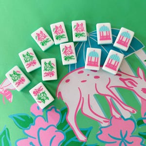 May include: White Mahjong tiles with pink and green floral and animal designs. The tiles are arranged on a green surface with a pink and white deer and floral pattern. The tiles feature a gazebo and a dog.