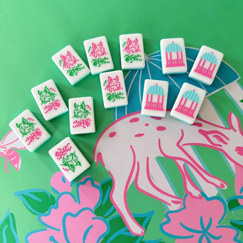 May include: White Mahjong tiles with pink and green floral and animal designs. The tiles are arranged on a green surface with a pink and white deer and floral pattern. The tiles feature a gazebo and a dog.