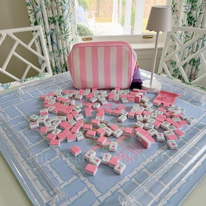 Pode incluir: Uma bolsa de cosméticos listrada em rosa e branco com um borla roxa está sobre uma superfície azul estampada. Numerosos azulejos de Mahjong rosa e branco estão espalhados. Uma pequena lâmpada branca com uma sombra ondulada também está presente.