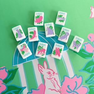 May include: A collection of white, rectangular game tiles with the word "Joker" printed on them. Each tile features a different pink and green animal or plant illustration. The tiles are arranged on a green and white patterned surface.