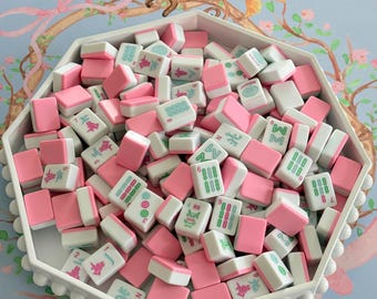 May include: A white, octagonal tray filled with pink and white Mahjong tiles. The tiles have green and blue symbols and numbers. The tray is on a light blue surface with a floral and animal design.
