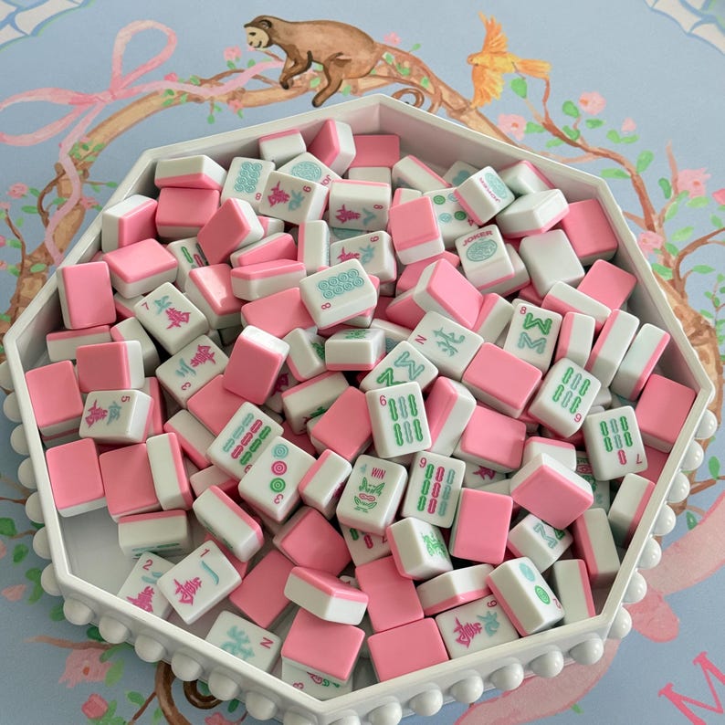 May include: A white, octagonal tray filled with pink and white Mahjong tiles. The tiles have green and blue symbols and numbers. The tray is on a light blue surface with a floral and animal design.
