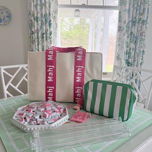May include: A cream-colored tote bag with pink straps and the word "Mahj" printed on them. A green and white striped cosmetic bag, a tray of Mahjong tiles, and a clear acrylic tile rack are also on the table. A small pink pouch is also visible.