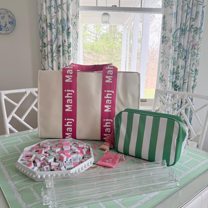 May include: A cream-colored tote bag with pink straps and the word "Mahj" printed on them. A green and white striped cosmetic bag, a tray of Mahjong tiles, and a clear acrylic tile rack are also on the table. A small pink pouch is also visible.
