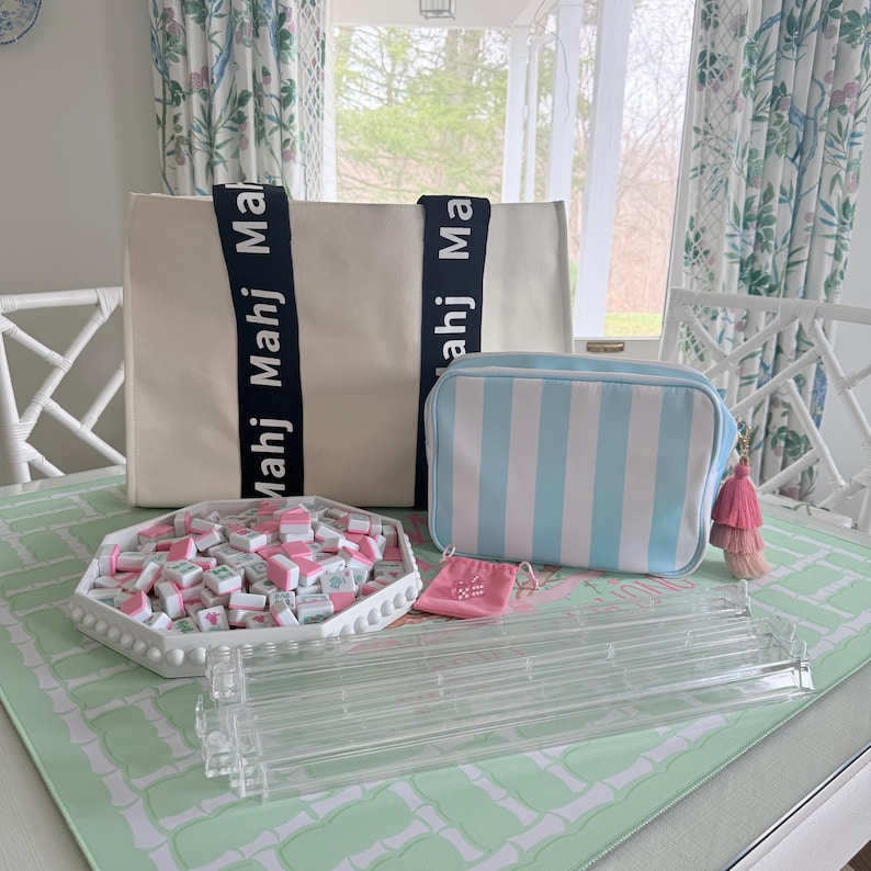 May include: A collection of Mahjong game accessories, including a cream-colored tote bag with navy blue straps, a striped blue and white cosmetic bag, and a white octagonal tray filled with pink and white tiles. A clear acrylic tile rack and a small pink pouch are also present.