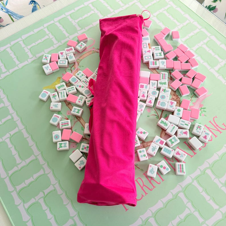 Puede incluir: Un estuche de transporte de tela rosa brillante colocado verticalmente en el centro, rodeado por una colecci&oacute;n dispersa de fichas de mahjong rectangulares. Las fichas presentan caras blancas con s&iacute;mbolos verdes y rojos.