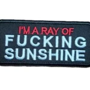 May include: Black rectangular patch with the text "I'M A RAY OF FUCKING SUNSHINE". The text is in white and red, with a black border. The patch is likely made of fabric and designed to be sewn or ironed onto clothing or accessories.