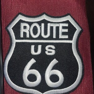 May include: A Route 66 patch with a black background and white border, featuring the words "ROUTE US 66" in white lettering. The patch is sewn onto a burgundy fabric.