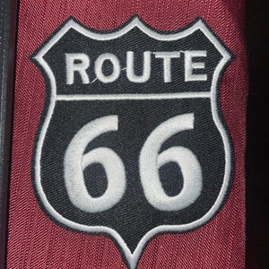 May include: Embroidered Route 66 patch in a shield shape. The patch features a black background with white text reading "ROUTE 66" and large white numbers "66". The patch is bordered with white stitching and set against a burgundy fabric.