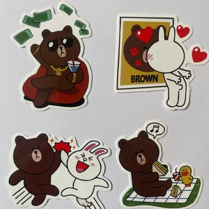 May include: Four cartoon stickers featuring a brown bear and a white rabbit. One sticker shows a bear with money and a drink. Another shows a bear and rabbit with hearts. The third shows the bear and rabbit high-fiving. The last shows a bear eating a sandwich.