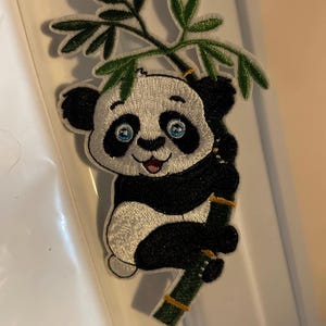 Könnte beinhalten: Gestickter Panda-Bären-Aufnäher mit schwarz-weißem Fell, blauen Augen und einem roten Mund. Der Panda hält einen grünen Bambusstiel und Blätter. Der Aufnäher ist wahrscheinlich für Kleidung oder Accessoires.