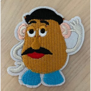 Puede incluir: Parche bordado con un personaje de dibujos animados con cabeza en forma de patata de color marrón, pelo negro, bigote negro y boca roja. El personaje tiene ojos blancos, orejas rosas y pies azules. El parche tiene un borde blanco y una mano saludando.