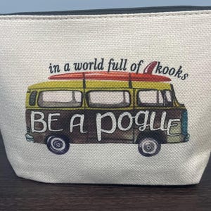Puede incluir: Bolsa de cosméticos de lona color crema con cremallera negra. La bolsa presenta una colorida ilustración de una furgoneta vintage con una tabla de surf y el texto "in a world full of kooks, BE A POGUE."