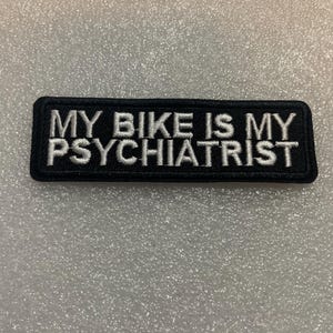 Pode incluir: Patch retangular preto com texto bordado branco que diz "MY BIKE IS MY PSYCHIATRIST". O patch tem uma borda preta e é projetado para ser costurado ou passado a ferro em roupas ou acessórios. Tema motociclista.