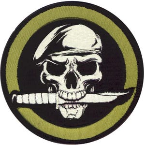 May include: Circular embroidered patch with a skull wearing a beret and a knife in its teeth. The design is in black and white, set against a black and olive green background. Suitable for application on clothing or accessories.