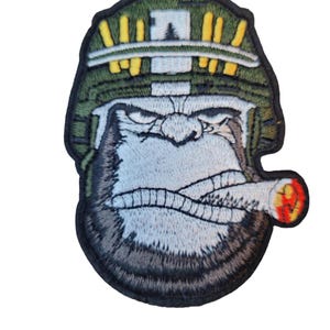May include: Embroidered patch featuring a gorilla wearing a military helmet and smoking a cigar. The patch has a black border, with the gorilla's face in shades of gray and brown. The helmet is green with yellow accents.