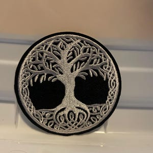 May include: A circular black patch featuring a white Celtic tree of life design. The tree's branches and roots are intricately detailed, with a border of interwoven patterns. This patch is suitable for clothing or accessories.