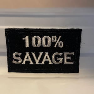 May include: Black rectangular patch with the words "100% SAVAGE" embroidered in silver thread. The patch is likely for clothing or accessories.
