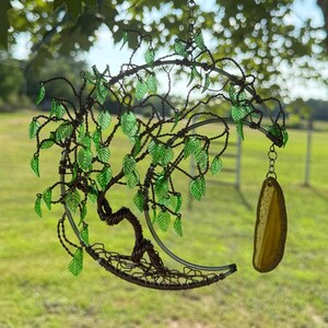 May include: A handcrafted metal tree sculpture with green glass leaves and a crescent moon shape. The sculpture features a twisted wire trunk and branches, with a yellow agate stone pendant hanging from the bottom. The sculpture is designed to be hung.