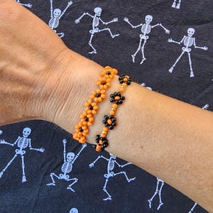May include: Two beaded bracelets. One is orange, the other is black with flower-shaped beads. The bracelets are on a wrist, against a black background with white skeleton designs. The bracelets are likely handmade.