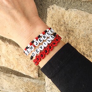 May include: Three beaded bracelets spelling out "NEW JERSEY DEVILS" in white letter beads. The bracelets are red, black, and white, with a floral design on the bottom bracelet. The bracelets are worn on a wrist.