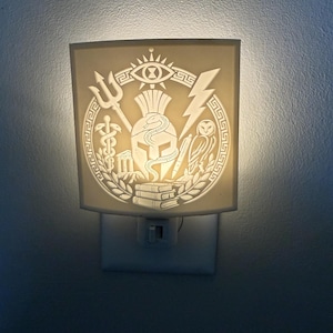 Könnte beinhalten: Ein weißes Steckdosen-Nachtlicht mit kreisförmigem Reliefdesign. Das Emblem zeigt einen zentralen Helm mit einer Schlange, flankiert von einem Dreizack, einem Blitz, einer Eule, einem Caduceus und Büchern, umrahmt von einem Mäander.