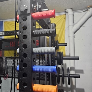 May include: A black weightlifting power rack with various colored grip handles. The handles are red, black, gray, blue, and orange. Weights are visible on the right side of the rack. Numbers are visible on the rack.