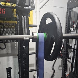 May include: A weightlifting barbell with a black weight plate and a blue resistance band attached. The barbell is mounted on a black weight rack. The image is taken indoors.