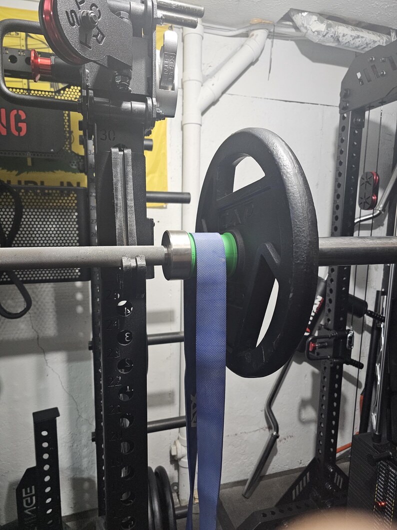 May include: A weightlifting barbell with a black weight plate and a blue resistance band attached. The barbell is mounted on a black weight rack. The image is taken indoors.