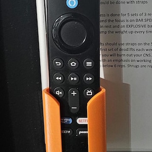 May include: Black Amazon Fire TV remote control in an orange holder. The remote has various buttons and the Amazon Alexa logo. The holder is mounted on a dark surface, with a workout plan visible in the background.