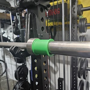 May include: A close-up of a weightlifting barbell with a green collar, secured in a black weight rack. The rack has multiple holes for adjustments and is surrounded by weight plates and other gym equipment. The word "YONG" is visible on the rack.