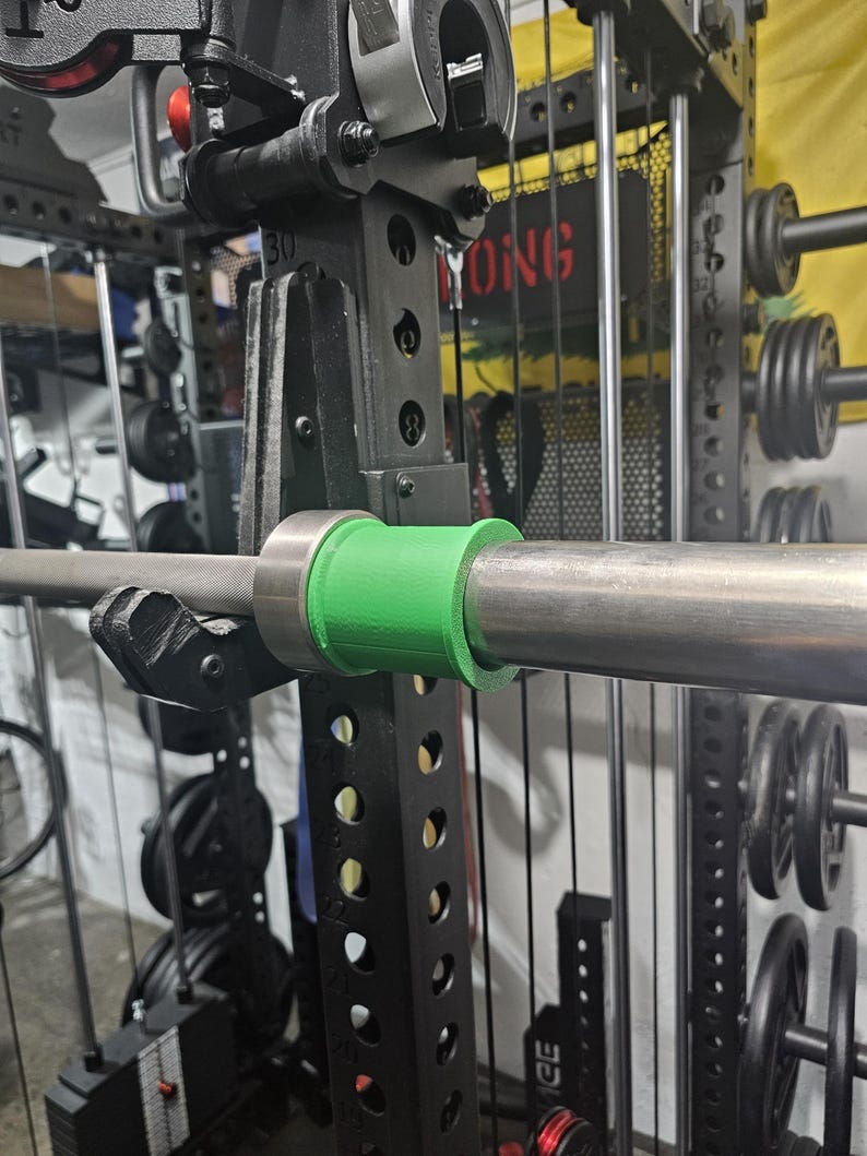 May include: A close-up of a weightlifting barbell with a green collar, secured in a black weight rack. The rack has multiple holes for adjustments and is surrounded by weight plates and other gym equipment. The word "YONG" is visible on the rack.