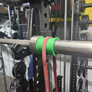 May include: A weightlifting setup featuring a barbell with green and red resistance bands attached. The barbell is secured within a black power rack, with weight plates visible in the background. The equipment is designed for strength training exercises.