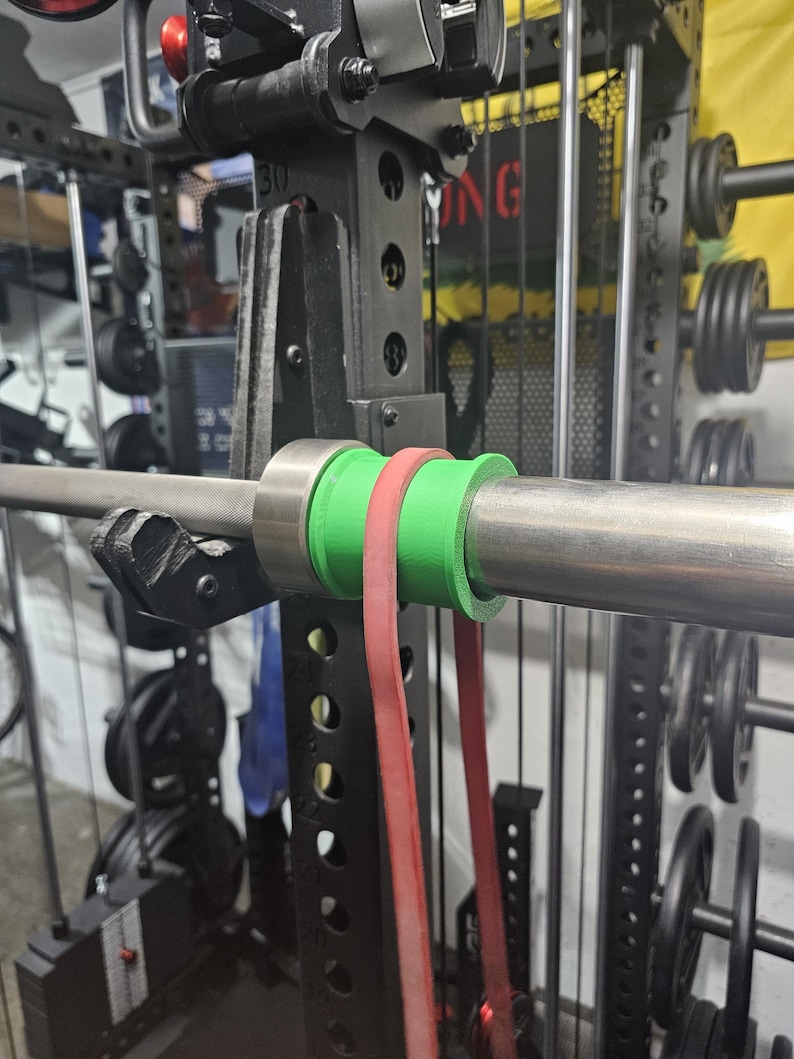 May include: A weightlifting setup featuring a barbell with green and red resistance bands attached. The barbell is secured within a black power rack, with weight plates visible in the background. The equipment is designed for strength training exercises.