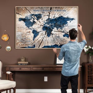 May include: A framed world map art print with a wood-grain background and a blue map design. The text "Home is Wherever I'm With You" is at the top, and "Adventures Thomas Family" is at the bottom. The print is being hung on a brown wall.