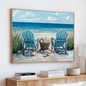 May include: A framed beach scene painting. Two light blue Adirondack chairs face the ocean, with a basket of seashells between them. The artwork depicts a sandy beach, blue ocean, and a bright sky with clouds. The frame is a light brown color.