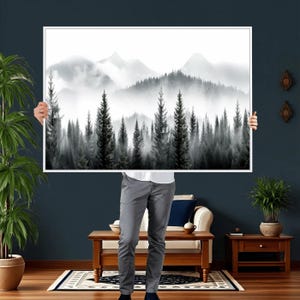 May include: A black and white framed print of a forest scene with tall evergreen trees and mountains in the background. The artwork is held up by a person in a room with a dark blue wall, a sofa, and wooden furniture.